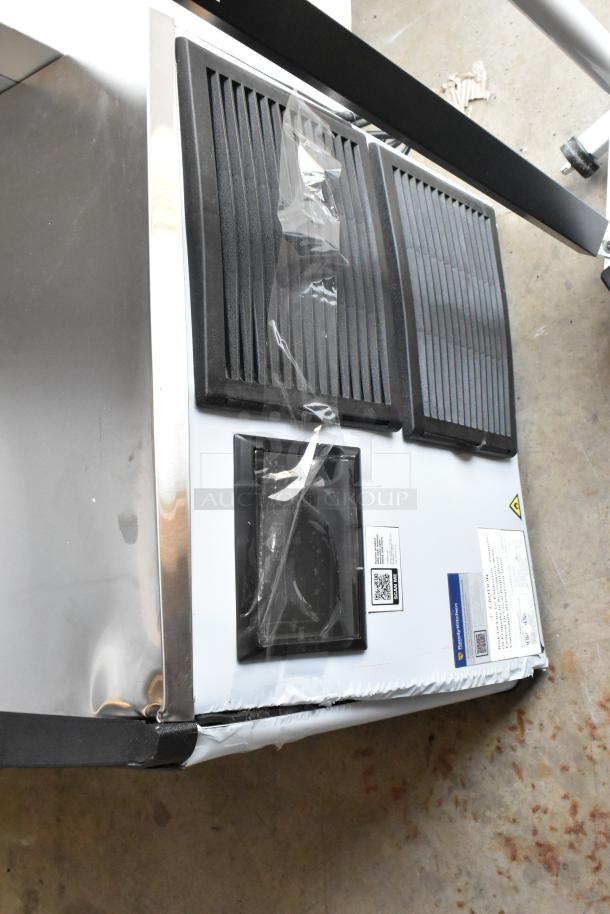 Commercial ice maker with black vents, stainless steel sides. Visible labels include QR code and caution notice. Slightly dented corner.