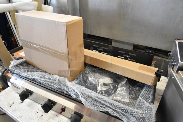 Commercial griddle with cardboard packaging, bubble wrap, and accessories on top. Looks new, with protective coverings and labels.