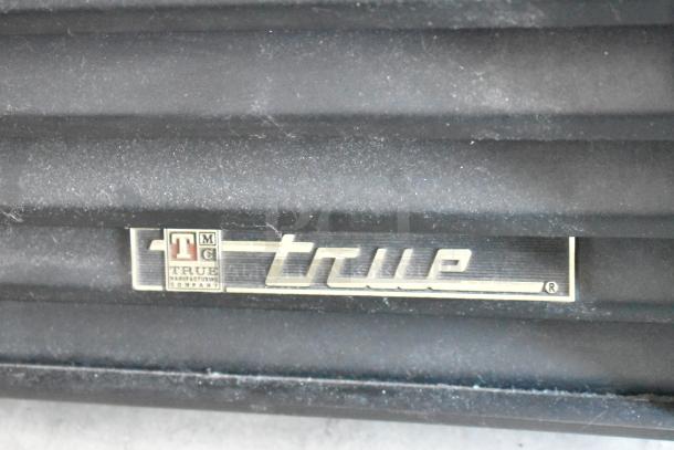 True Manufacturing Company logo on black appliance, features chrome lettering and emblem. Condition: light dust present.