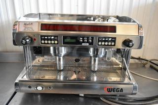 Wega espresso machine with dual group heads, stainless steel finish, programmable buttons, visible wear, branded logo.
