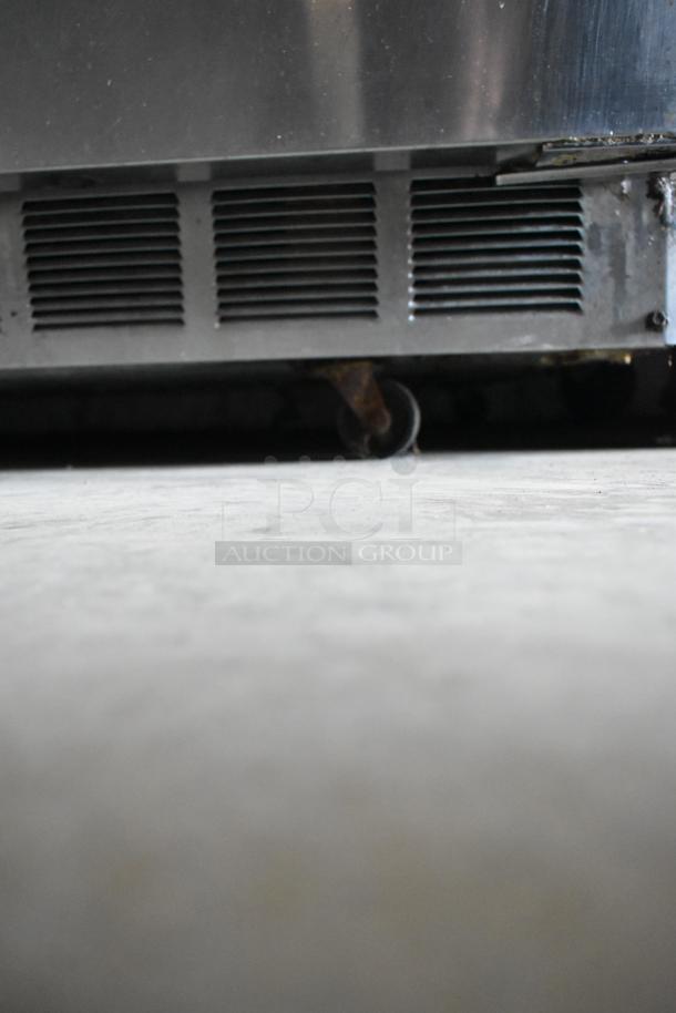 Industrial metal refrigerator base with visible vents and a caster wheel, showing signs of wear.