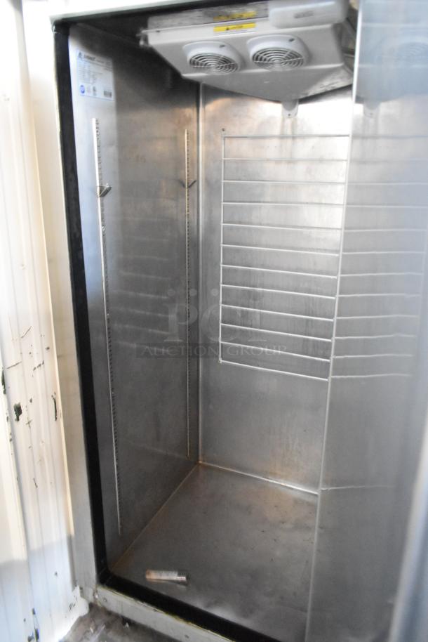Commercial stainless steel refrigerator, interior view, ventilated top, adjustable shelves, visible wear, NSF label.