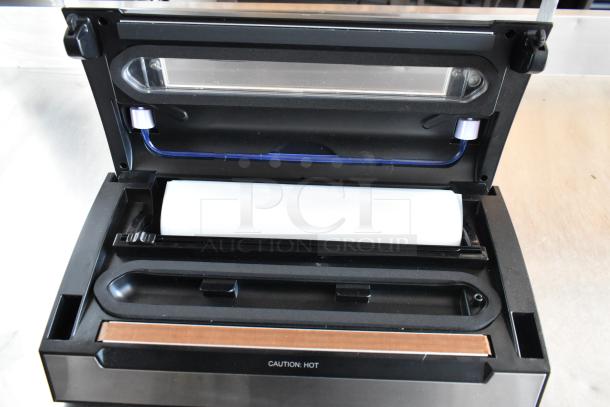 Open black vacuum sealer machine with roll compartment, "CAUTION: HOT" label inside. Appears in good condition.
