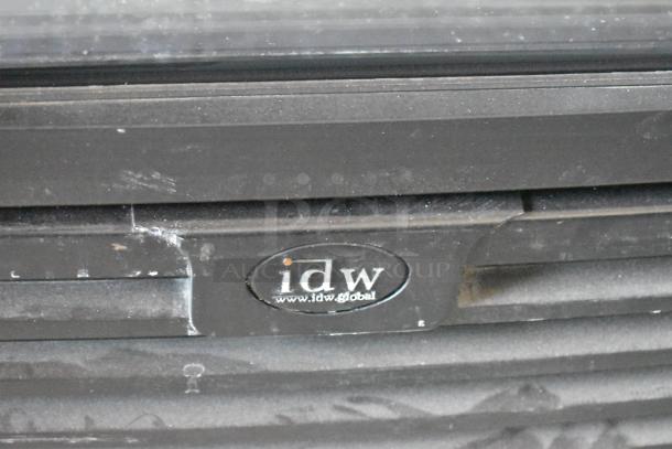 Close-up of a black appliance with an "idw" logo. Shows signs of wear and surface scratches. Logo indicates idwglobal website.