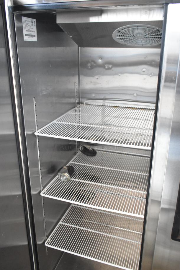 Commercial stainless steel refrigerator interior with four wire shelves, ventilation fan, and visible manufacturing label.