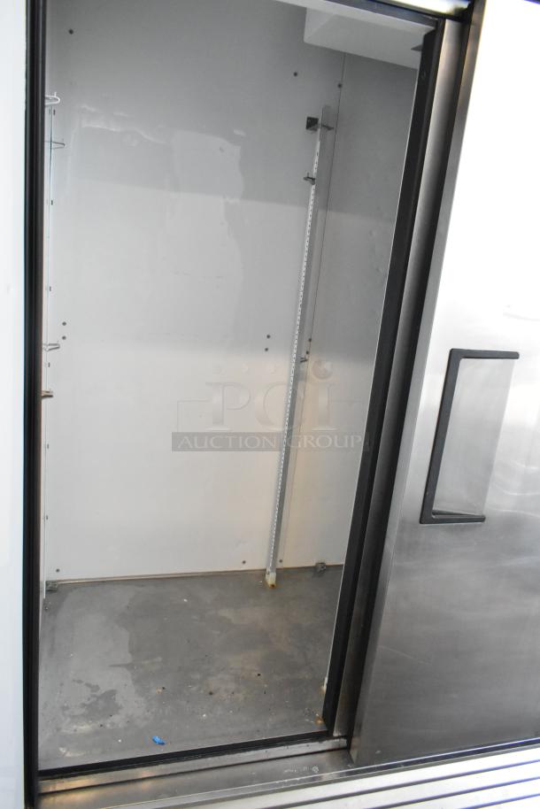 Commercial stainless steel refrigerator with a sliding door, empty interior, adjustable shelving slots, and minor floor wear.
