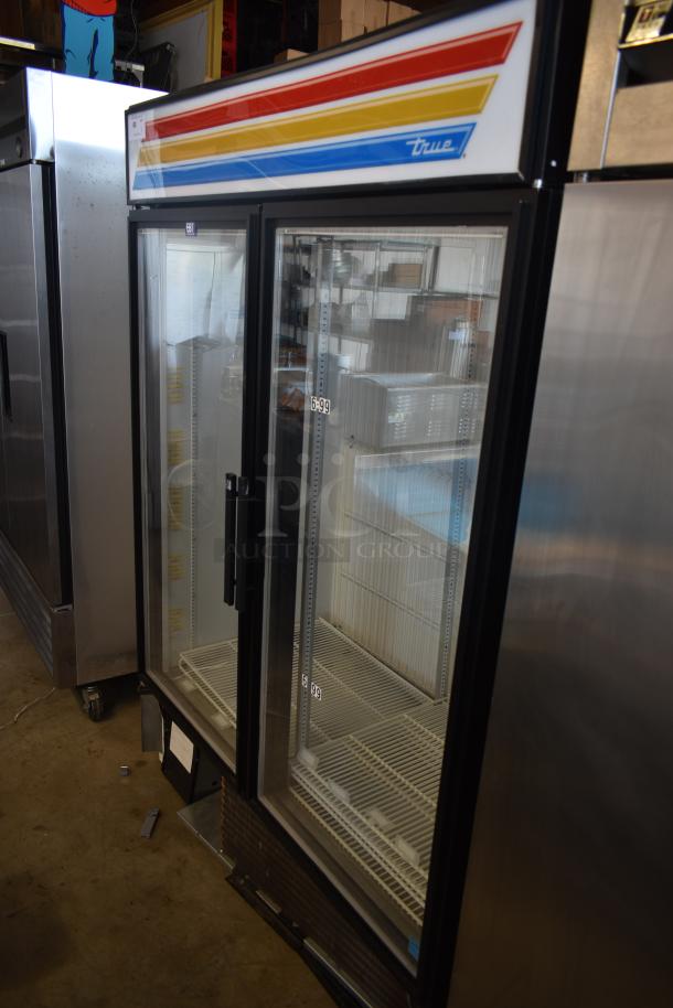 True commercial glass door refrigerator with colorful top panel. Features adjustable shelves, visible serial number, and sturdy handle.