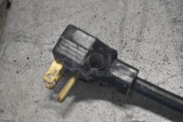 Black electrical plug with visible wear, featuring two brass prongs against a gray concrete background.