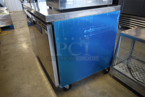 Commercial stainless steel undercounter refrigerator on wheels, blue protective film partially intact, minor scuffs visible.