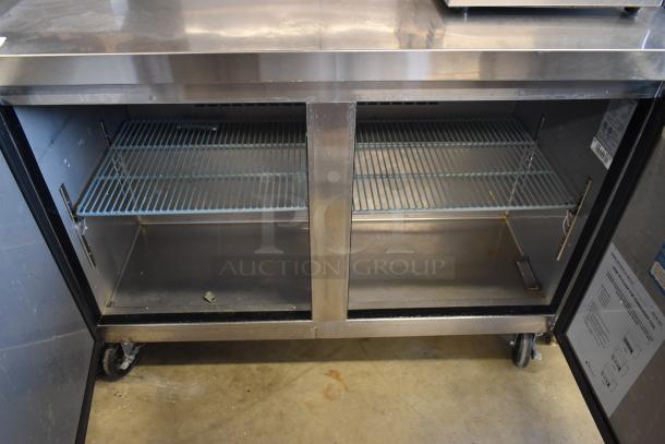 Stainless steel commercial under-counter refrigerator with two wire shelves, wheels, and open doors. Minor wear visible.