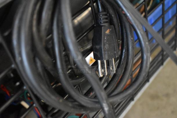 Black extension cord with three-prong plug, coiled on a wire rack, showing slight wear and small rust spots.