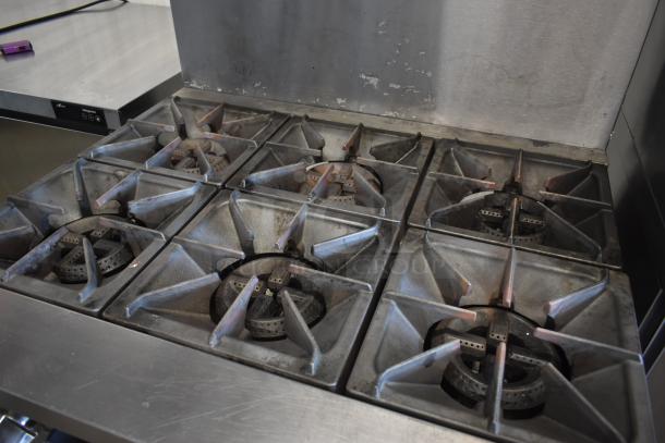 Commercial six-burner gas stove with cast iron grates, showing signs of use. Stainless steel surface and backsplash.