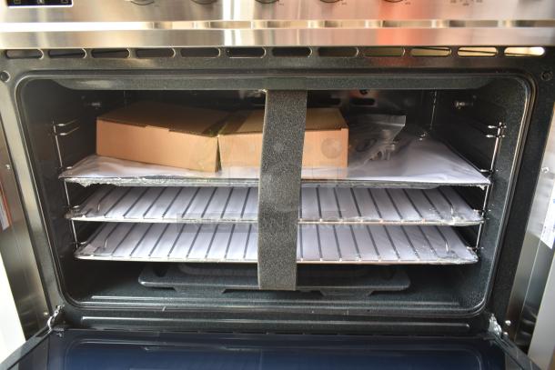 Stainless steel oven with open door showing racks, packaging, and shelving material. New condition, interior stickers visible.