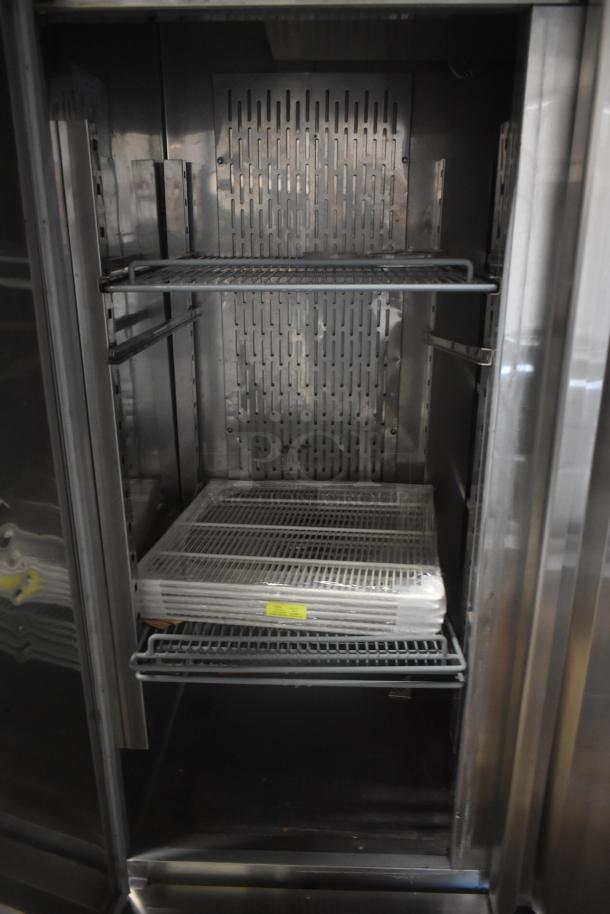 Commercial stainless steel refrigerator, open with wire racks, shows wear. Includes wrapped extra shelves with visible yellow label.