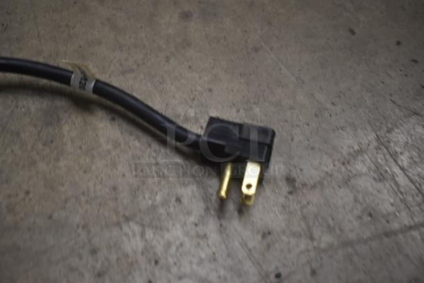 Close-up of a black electrical plug with three brass prongs, attached to a black cable. A small label reads "FR." in good condition.