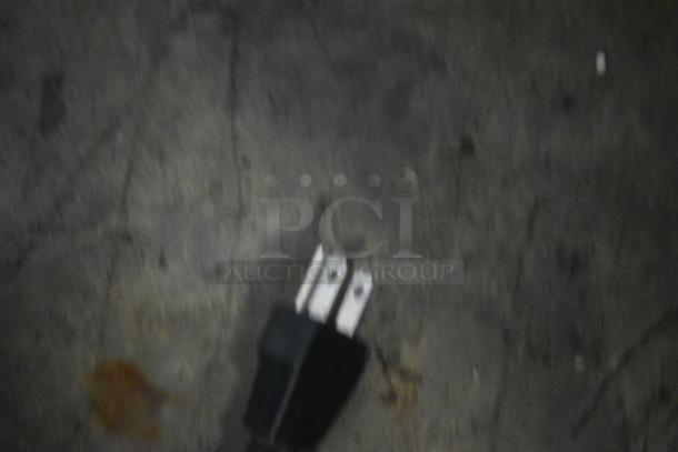 Blurry image of a two-prong electrical plug on a dark surface. No visible identifying marks or labels.