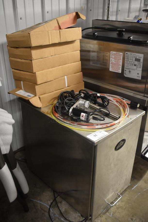 Industrial beverage dispenser with wiring and hoses on top, stacked cardboard boxes, labels showing operating instructions and safety warnings.
