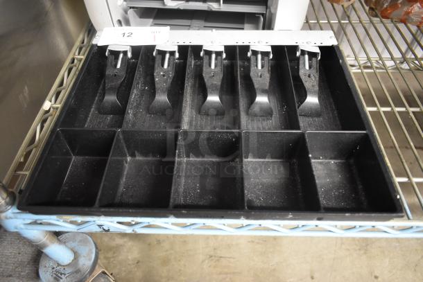 Black cash register drawer with six compartments and metal clips. Shows organizational slots for bills and coins, minor wear.