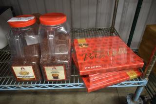 Two large containers of chocolate sprinkles with red lids next to Hershey's Pot of Gold chocolate boxes on a metal shelf.