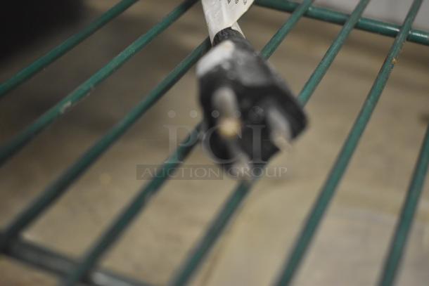 Blurred close-up of a two-prong plug with a label attached, placed on a green wire rack.