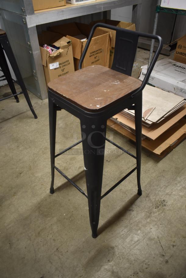 Industrial-style black metal bar stool with wooden seat, showing signs of wear. Square backrest and sturdy legs.