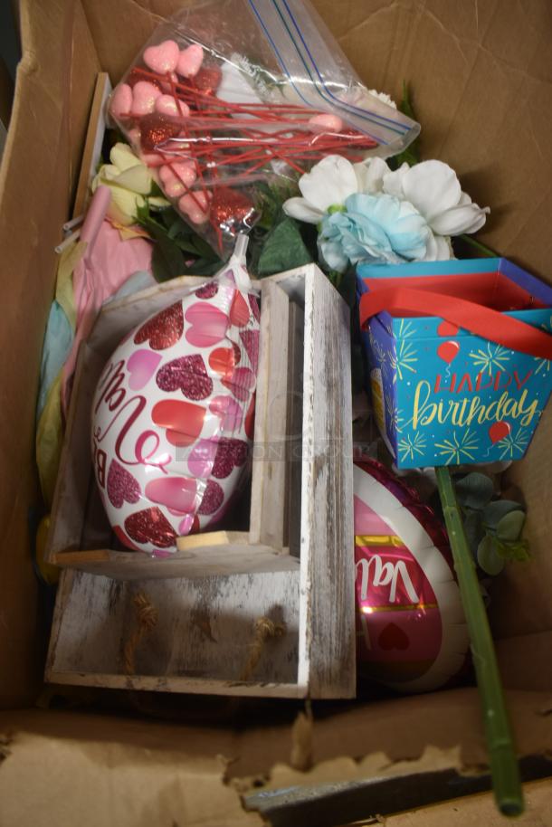 Cardboard box with mixed party items including balloons, gift bags, faux flowers, and heart decorations.