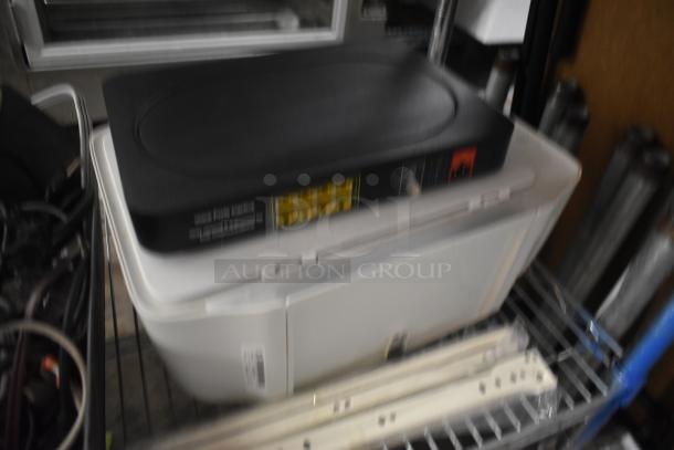 Blurry image of an electronic device on a metal shelf, possibly a printer or scanner, with visible labels and markings.