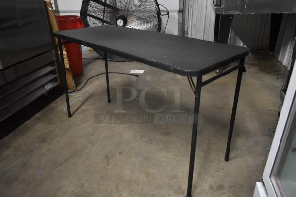 Black folding table, rectangular shape, metal legs, minor surface wear, suitable for indoor or outdoor use.