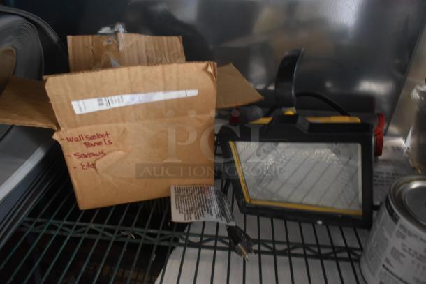 Box labeled "Wall Socket Panels," "Screws," and "Etc," next to a portable work light with cord and plug.
