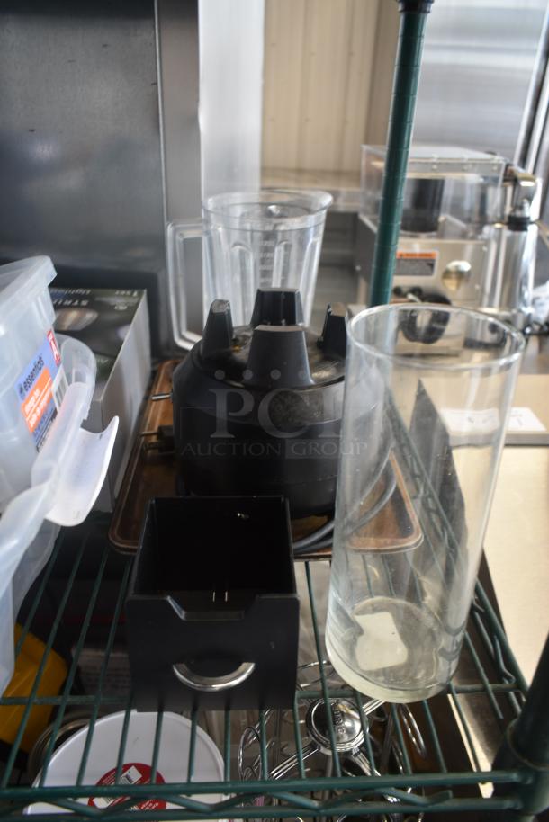 Commercial blender base with two clear blender jars, used condition, on a metal shelf. Power cord visible.