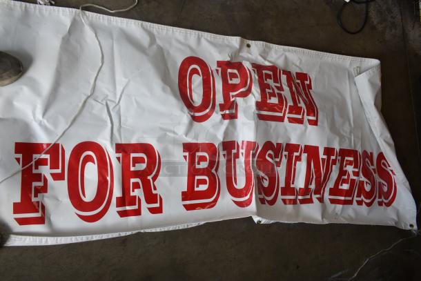 Large white vinyl banner with red "OPEN FOR BUSINESS" text, grommets for hanging, showing wrinkles and light wear.