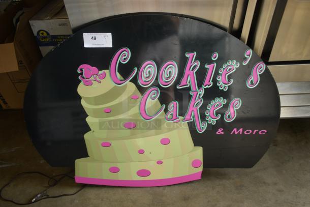 Decorative sign for "Cookie's Cakes & More" with a cake graphic, pink and green text on black, label "49" visible.
