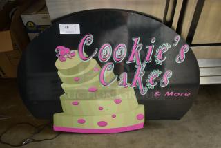 Decorative sign for "Cookie's Cakes & More" with a cake graphic, pink and green text on black, label "49" visible.