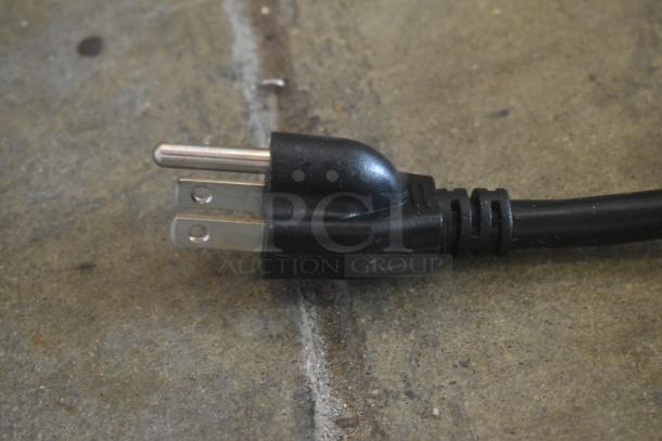 Black electrical power plug with two flat prongs and one round prong, in used condition on a concrete surface.