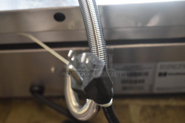 Close-up of a heavy-duty appliance plug with a metal sheath cable, attached to a metal surface. Label with barcode visible in the background.