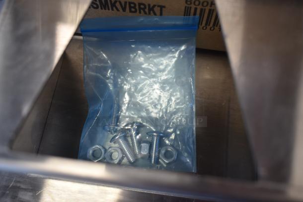 Bag of screws and nuts in a clear resealable bag, viewed inside a metal container. Label reads "SMKVBRKT 600A."