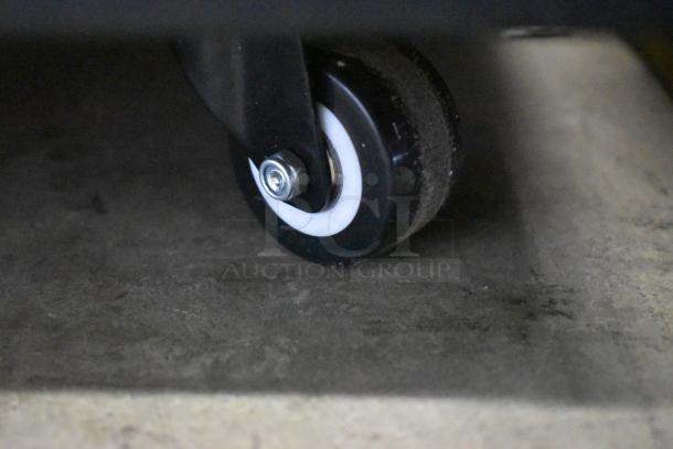 Black caster wheel on concrete floor, attached to metal frame, featuring white ring and metal bolt. Appears in good condition.
