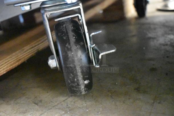 Close-up of a metal caster wheel with a brake, mounted on a wooden structure. The wheel shows slight wear.