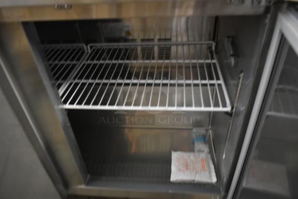 Commercial stainless steel refrigerator, interior view showing two shelves and a packet of desiccant. Clean and well-maintained.