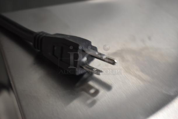 Black electrical power plug with two metal prongs, slight wear, resting on a metallic surface.