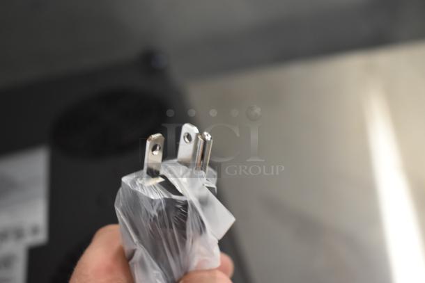 Close-up of a wrapped electrical plug with three prongs, indicating new condition. Background shows part of the appliance with a vent.