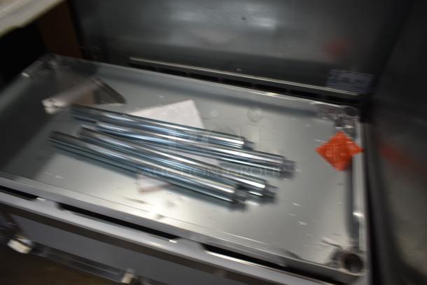Metal rods on a metal tray, possibly part of industrial equipment. Includes red tag and paperwork, indicating unused condition.