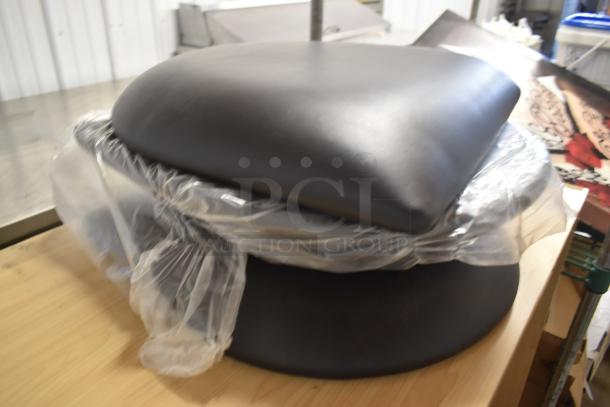 Black leather chair cushion set, new condition, partially wrapped in plastic, on a wooden surface.