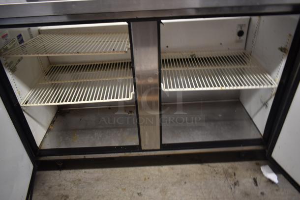 Commercial stainless steel undercounter refrigerator with two shelving units. Visible wear, no visible branding.