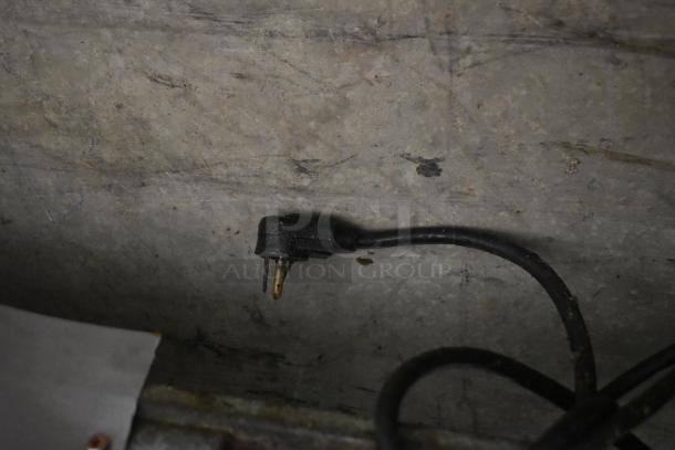 Black electrical plug with two prongs, attached to a black cord. Plug shows signs of wear and some corrosion.