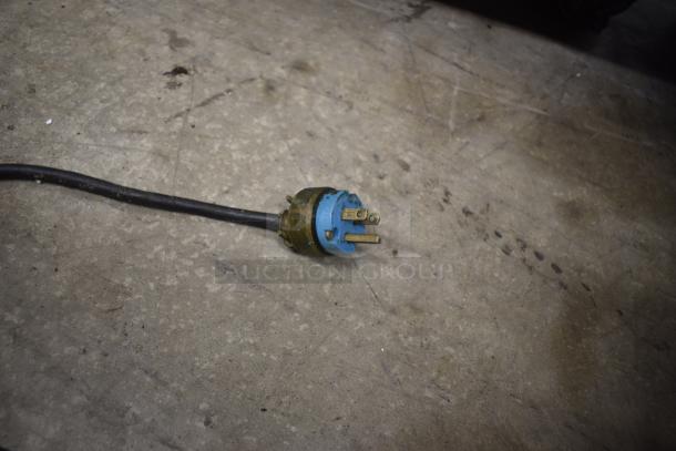 Blue and brass three-prong electrical plug with black cable, worn condition, on a concrete floor.