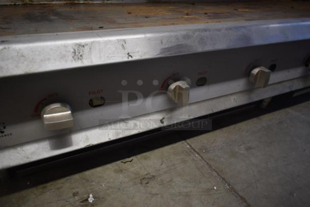 Stainless steel commercial griddle with control knobs, showing signs of wear. Markings indicate ON/OFF and pilot settings.