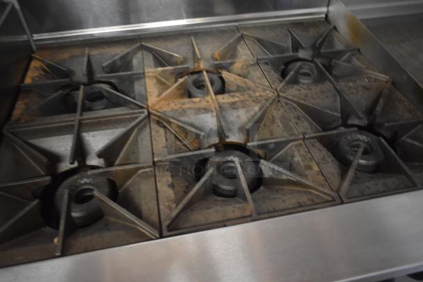 Commercial six-burner gas stovetop, used condition with visible wear. Ideal for restaurant kitchens. Metal grates.