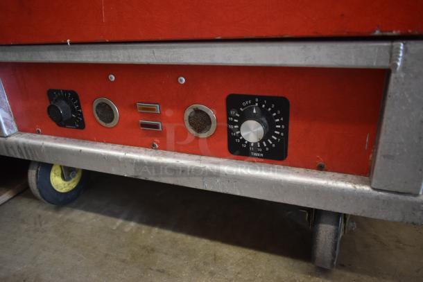 Red industrial machine base with metal edges, featuring dials for thermostat and timer settings, wear visible. Mounted on wheels.
