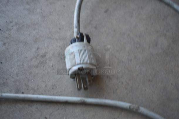 Blurry image of an electrical plug with three prongs, connected to a gray cable. Condition appears worn. No visible markings.